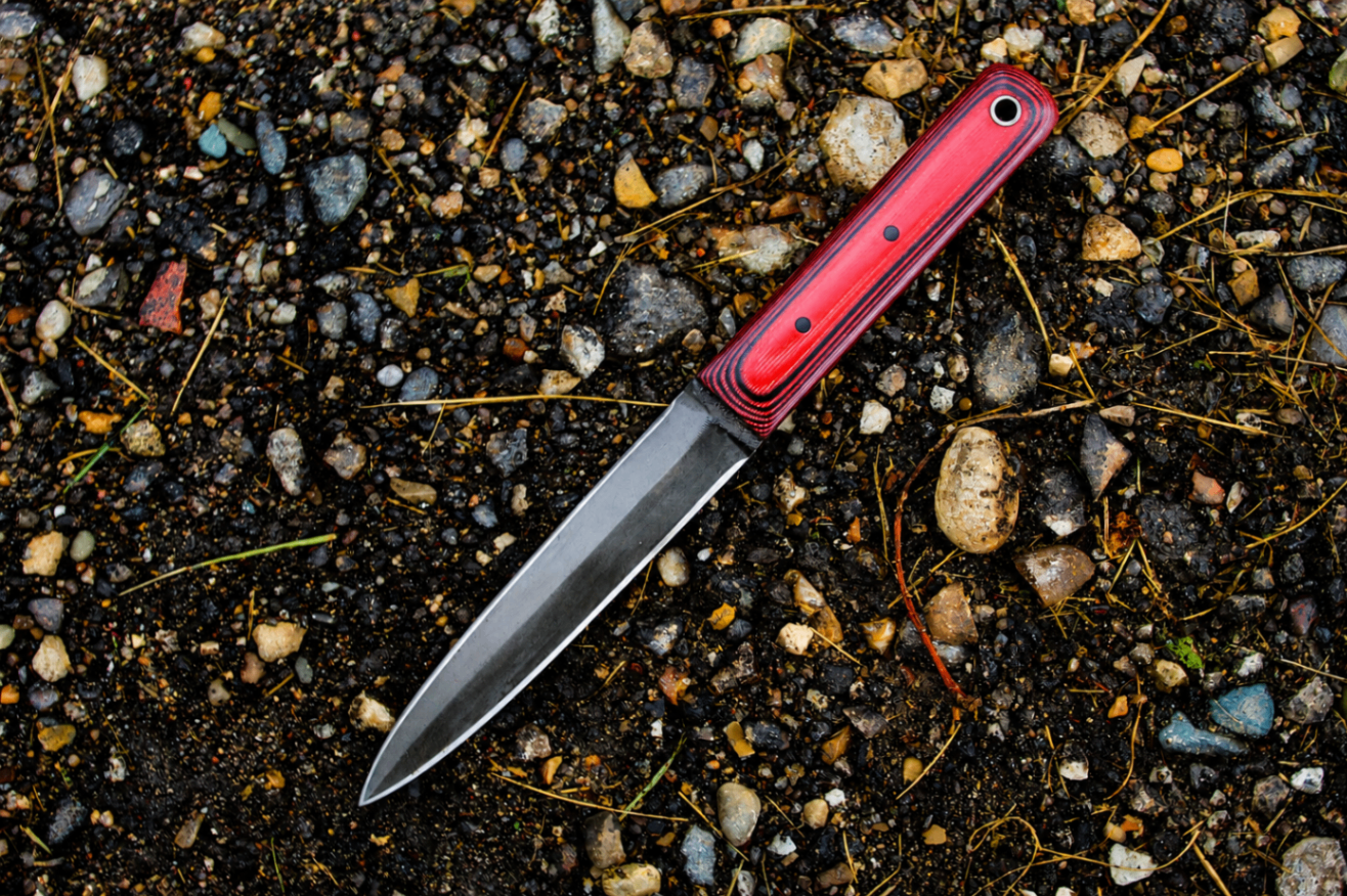 Handcrafted dagger with red micarta handle resting on gravel and earth