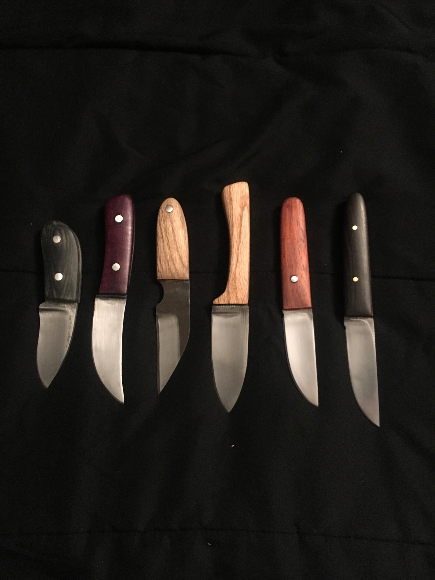 Lineup of six handcrafted knives with varied wood handles on black fabric