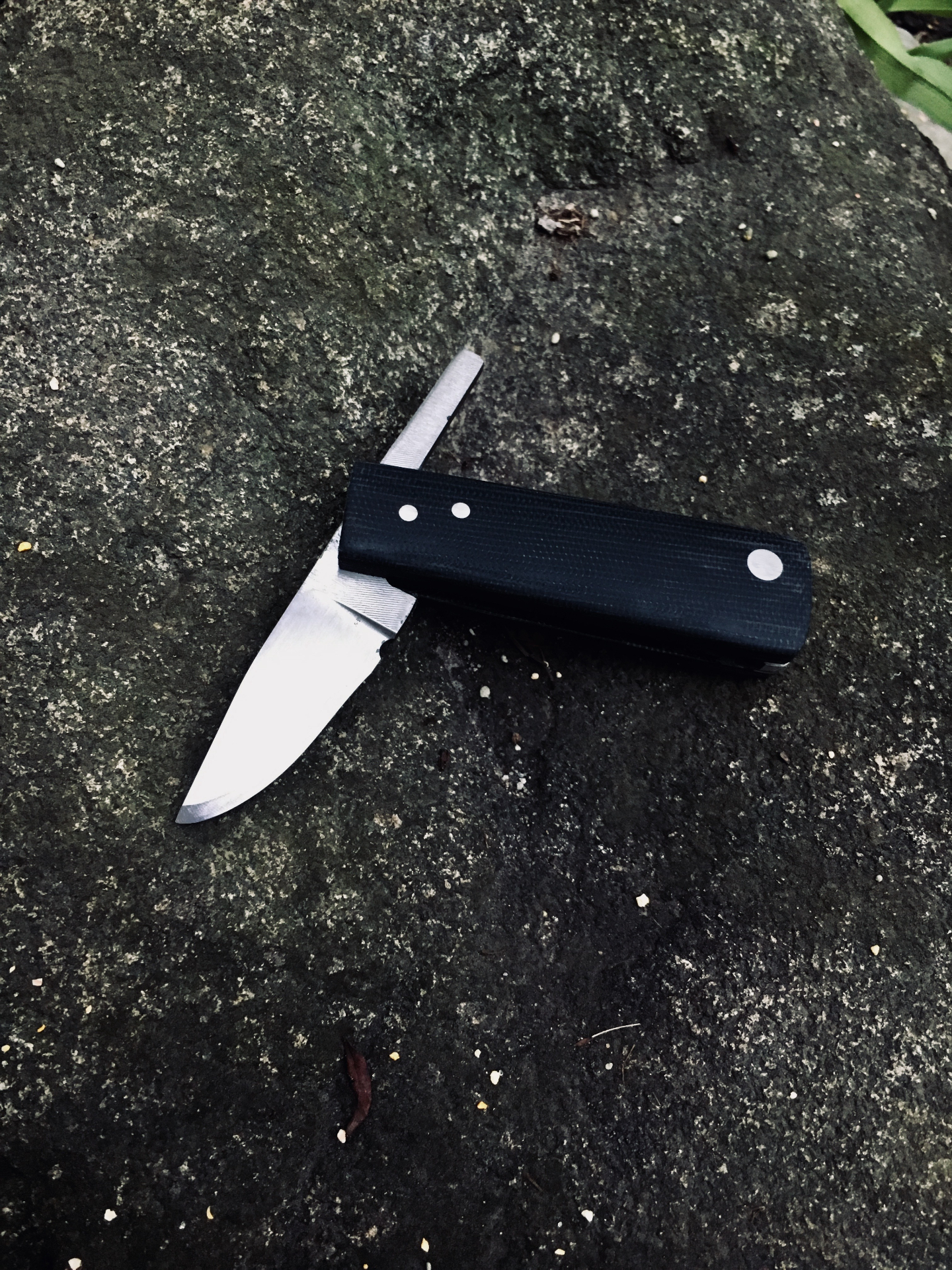 Handcrafted slip joint folding knife with black G10 handle resting on a mossy stone